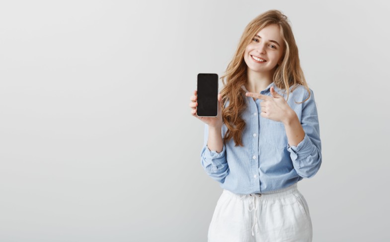 super-useful-device-pleased-good-looking-female-student-with-blond-hair-blue-collar-shirt-showing-black-smarpthone-pointing-gadget-with-index-finger-offering-buy-item-min